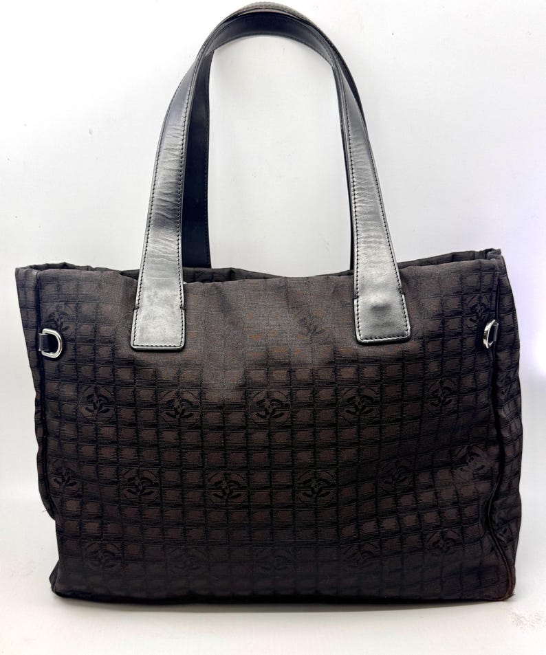 K&ouml;nnte beinhalten: Dunkelbraune Tragetasche mit sich wiederholendem Muster. Die Tasche hat zwei schwarze Ledergriffe und silberfarbene Metallringe an den Seiten. Das Material der Tasche hat einen dezenten Glanz.