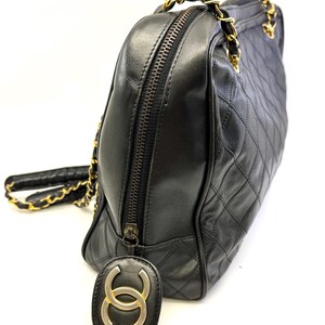 May include: Black quilted leather handbag with gold-tone chain straps and zipper closure. The bag features a diamond pattern and a gold and black logo charm. The bag is a classic style.