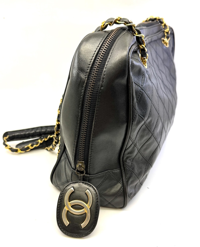 May include: Black quilted leather handbag with gold-tone chain straps and zipper closure. The bag features a diamond pattern and a gold and black logo charm. The bag is a classic style.