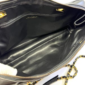 May include: Black leather handbag with gold-tone chain strap and hardware. The interior features a black lining and a zippered pocket. The brand name "CHANEL" is visible inside the bag. The bag has a rectangular shape and a top zipper closure.