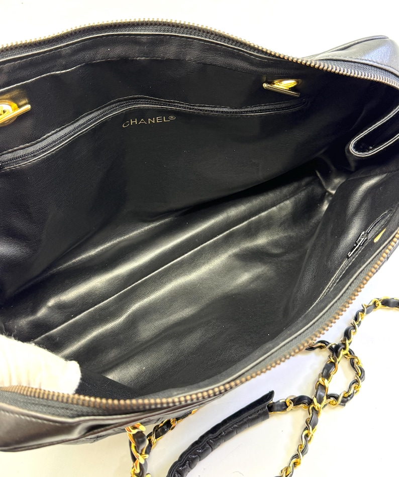 May include: Black leather handbag with gold-tone chain strap and hardware. The interior features a black lining and a zippered pocket. The brand name "CHANEL" is visible inside the bag. The bag has a rectangular shape and a top zipper closure.