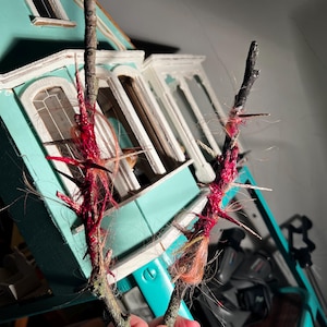 May include: Two small, textured branches with red and tan accents. The branches are held in front of a miniature teal house with white trim. The branches have a unique, handmade appearance.