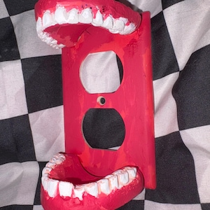May include: A red painted electrical outlet cover with a set of teeth attached at the top and bottom. The teeth are white with pink gums. The background is a black and white checkered pattern.