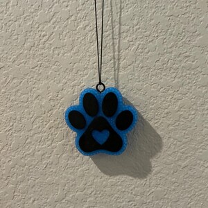 May include: A blue and black paw print-shaped ornament with a heart in the center. The ornament is hanging from a black string. The paw print has a glittery texture and is against a textured white wall.