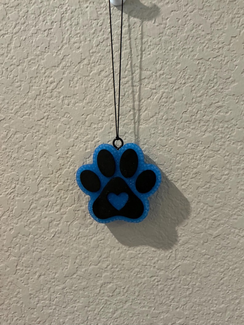 May include: A blue and black paw print-shaped ornament with a heart in the center. The ornament is hanging from a black string. The paw print has a glittery texture and is against a textured white wall.
