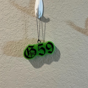 Pu&ograve; includere: Un ornamento sospeso verde neon brillante con il testo nero 659 in un carattere in stile gotico. L'ornamento &egrave; appeso a un cordino nero sottile fissato a un gancio adesivo bianco. L'oggetto presenta un aspetto testurizzato e semitrasparente.