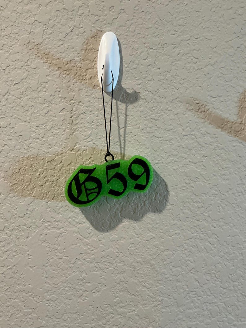 Pu&ograve; includere: Un ornamento sospeso verde neon brillante con il testo nero 659 in un carattere in stile gotico. L'ornamento &egrave; appeso a un cordino nero sottile fissato a un gancio adesivo bianco. L'oggetto presenta un aspetto testurizzato e semitrasparente.