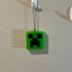 May include: A square, bright green, translucent resin ornament featuring a black pixelated face design. The ornament is suspended by a thin black cord from a white plastic adhesive hook. The surface of the green resin has a subtle glittery texture.