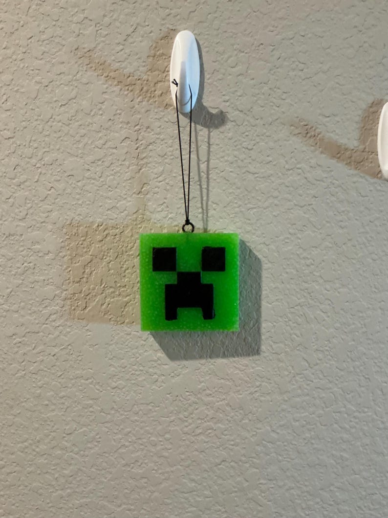 May include: A square, bright green, translucent resin ornament featuring a black pixelated face design. The ornament is suspended by a thin black cord from a white plastic adhesive hook. The surface of the green resin has a subtle glittery texture.