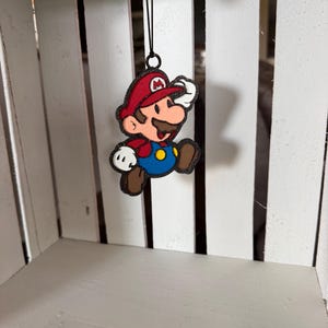 May include: A hanging ornament featuring a glittery, two-dimensional depiction of the character Mario. The figure wears a red cap with a white M, a red shirt, blue overalls with yellow buttons, white gloves, and brown shoes, suspended by a black string.