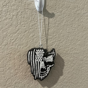 May include: Black and white air freshener in the shape of a skull with an American flag design. The air freshener is hanging from a white hook on a textured wall.
