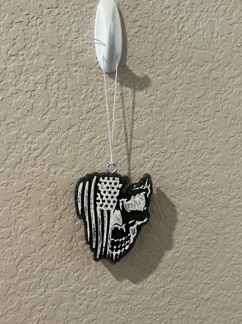 May include: Black and white air freshener in the shape of a skull with an American flag design. The air freshener is hanging from a white hook on a textured wall.