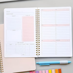 Peut inclure: Un agenda à spirales ouvert, avec une spirale dorée. La page de gauche comporte des sections pour le focus hebdomadaire, les priorités, la liste des tâches, les notes et un suivi des habitudes. La page de droite est pour la planification quotidienne.