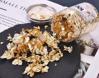 Può includere: Un barattolo trasparente di scaglie di foglia d'oro, con alcune sparse su una superficie nera. Il barattolo è etichettato "10g" e "Gold for food". Il coperchio è aperto e all'interno c'è della foglia d'oro.