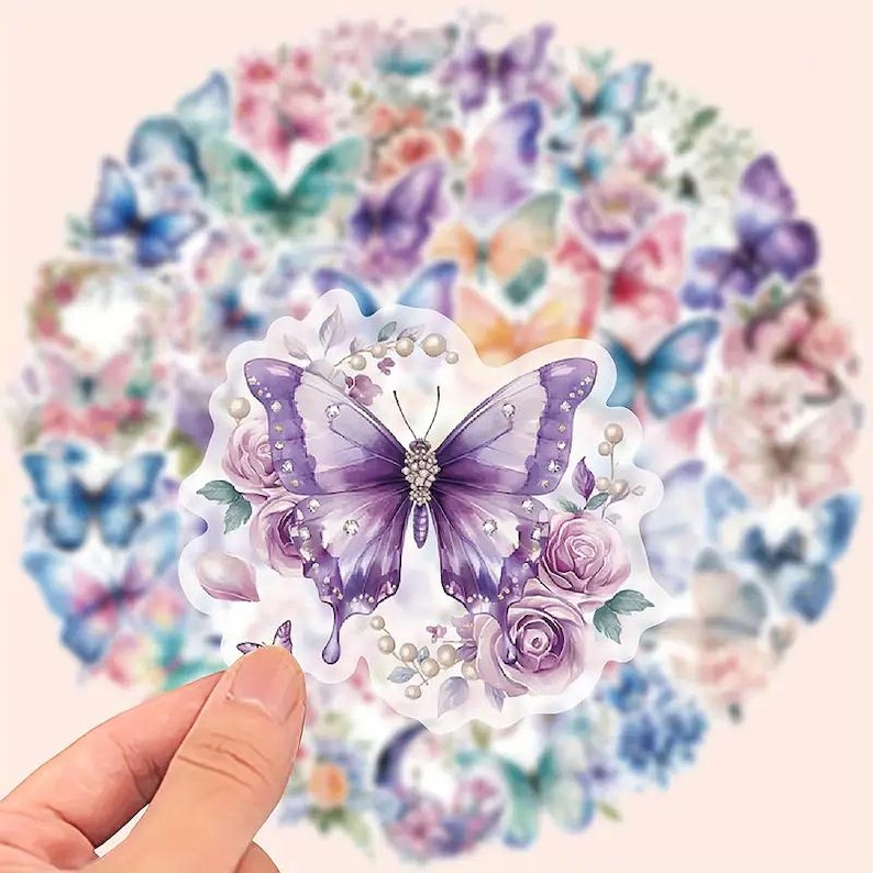 May include: A collection of butterfly stickers in various colors, including purple, blue, and pink. The stickers feature detailed butterfly designs with floral accents. The stickers are held by a hand, with a soft pink background.