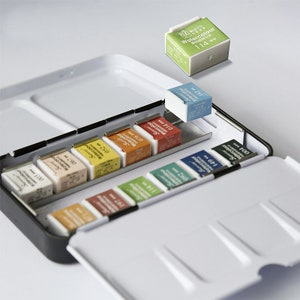 Puede incluir: Una caja de metal abierta con un interior blanco que contiene un juego de 14 pastillas de pintura de acuarela. Las pastillas están dispuestas en filas y están etiquetadas con números. La caja está abierta y la tapa está levantada. La tapa tiene un fondo blanco con un borde negro. Las pastillas de pintura son de varios colores, incluyendo rojo, naranja, amarillo, verde, azul y morado. La caja está etiquetada con "Watercolour 114" en la tapa.