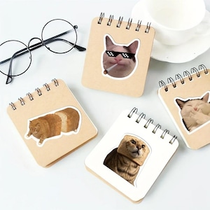 Op de afbeelding: Een verzameling spiraalgebonden notitieboekjes met kat-thema stickers. De stickers tonen verschillende kattenmemes, waaronder een kat met een zonnebril en een sigaret, en een kat met brood om zijn hoofd. Ronde brillen zijn ook inbegrepen.