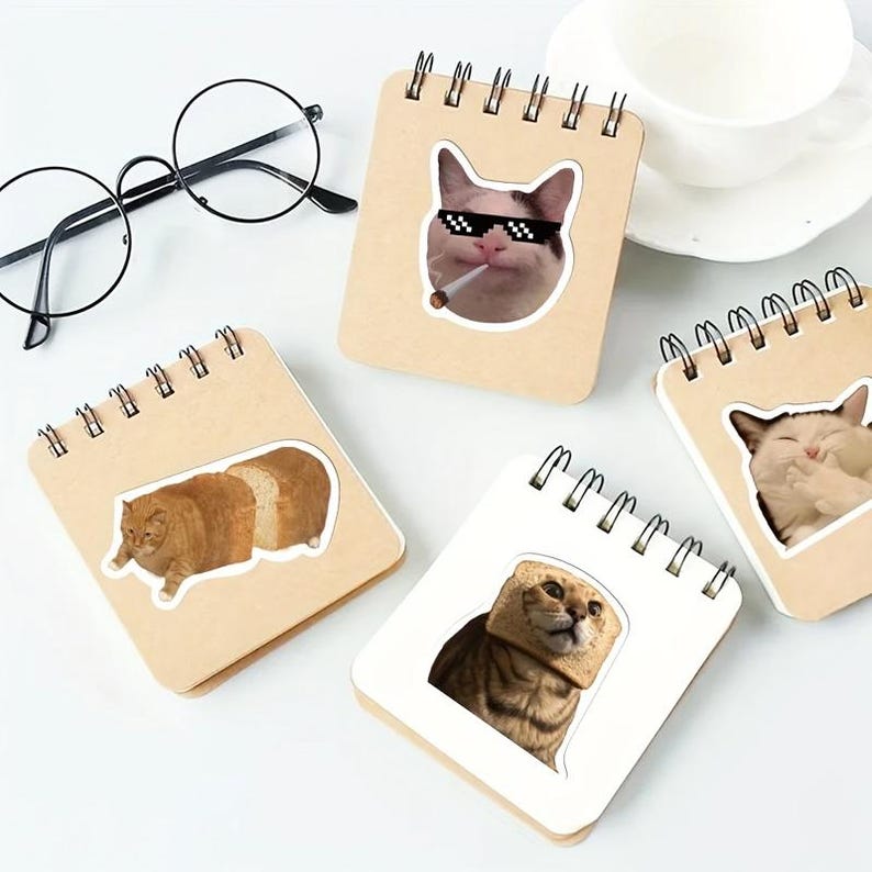 Op de afbeelding: Een verzameling spiraalgebonden notitieboekjes met kat-thema stickers. De stickers tonen verschillende kattenmemes, waaronder een kat met een zonnebril en een sigaret, en een kat met brood om zijn hoofd. Ronde brillen zijn ook inbegrepen.