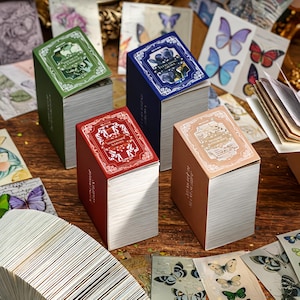 May include: Four small stacks of paper with decorative covers in green, blue, red, and tan. The covers feature ornate designs and text. Butterfly-themed stationery is scattered around the stacks, with various butterfly illustrations.