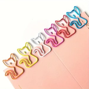 Könnte beinhalten: Eine Sammlung von katzenförmigen Büroklammern in den Farben Orange, Gelb, Weiß, Rosa, Rot und Blau. Die Klammern sind diagonal auf einem rosa Notizblock angeordnet. Diese dekorativen Klammern verleihen jeder Büro- oder Schule eine spielerische Note.