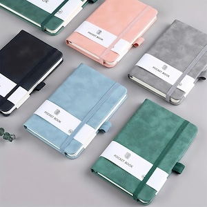 Può includere: Una collezione di taccuini tascabili colorati, in nero, rosa, grigio, blu e verde. Ogni taccuino presenta un elastico abbinato e una fascia di carta bianca con la scritta "POCKET BOOK".