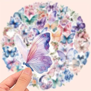 May include: A hand holding a translucent butterfly sticker with watercolor-style wings in shades of purple, pink, and blue. The background features a blurred collection of butterfly stickers and floral designs in various colors.