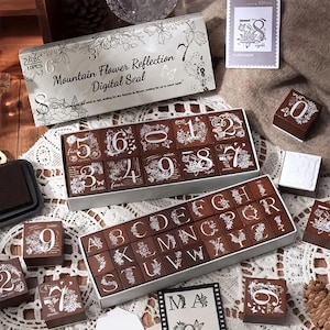 Könnte beinhalten: Eine Sammlung von Holzstempeln mit weißen floralen Designs sowie Zahlen und Buchstaben. Die Stempel befinden sich in einer Box mit dem Text "Mountain Flower Reflection Digital Seal". Einzelne Stempel und ein Stempelkissen sind ebenfalls vorhanden.