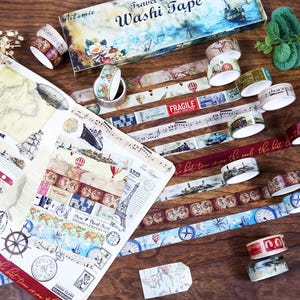 May include: A collection of travel-themed washi tape rolls and a box. The tape features designs of maps, hot air balloons, ships, and musical notes. The box is decorated with a nautical scene and the words "Travel Washi Tape".