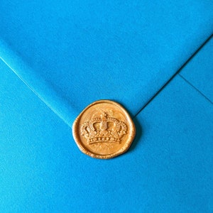 May include: A gold wax seal with a detailed crown design is affixed to a bright blue envelope. The seal is round and has a textured surface, adding a touch of elegance. The envelope's color is a vibrant, solid blue.