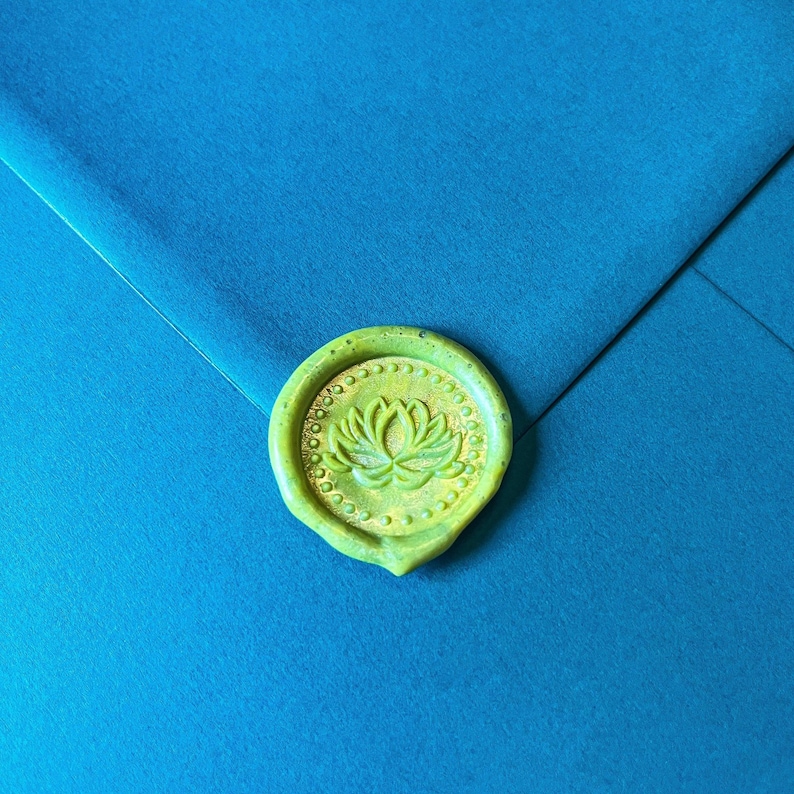 May include: A close-up of a teal envelope sealed with a green wax stamp. The stamp features a detailed lotus flower design surrounded by a dotted border. The wax seal is a vibrant green color, contrasting with the teal envelope.