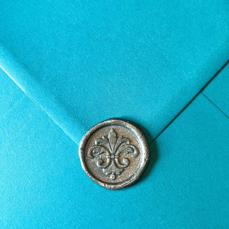 May include: Close-up of a teal envelope sealed with a silver wax stamp. The stamp features a fleur-de-lis design. The envelope is folded, creating diagonal lines across the surface. The wax seal adds a touch of elegance.