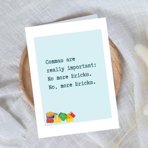 May include: A white greeting card with a light blue background and the text "Commas are really important: No more Bricks. No, more Bricks." Colourful toy bricks are at the bottom of the card. The card is on a wooden plate.