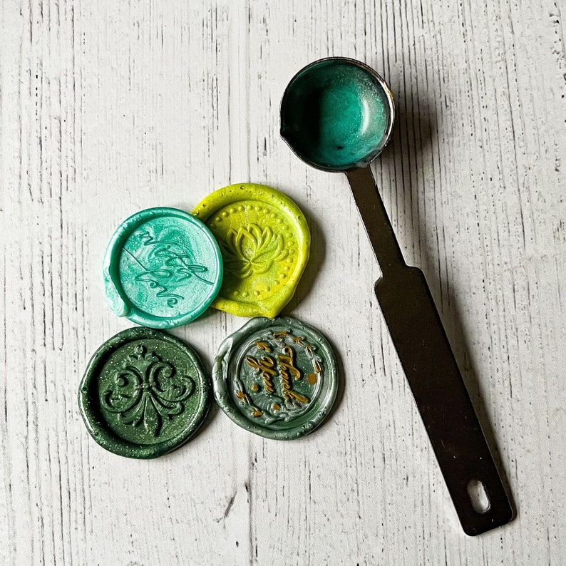 May include: A collection of colorful wax seals with various designs, including a fleur-de-lis and a lotus flower. A teal-colored wax is in a metal melting spoon. The seals are in shades of green and yellow, with gold accents. The background is a white wooden surface.