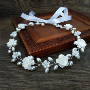 May include: A white floral crown with pearl accents. The crown is made of white flowers and pearls on a silver wire base. The crown is lying on a wooden surface.