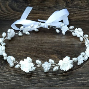 May include: A white floral crown with pearl accents and a white satin ribbon. The crown is made of wire and is decorated with small white flowers and pearls.