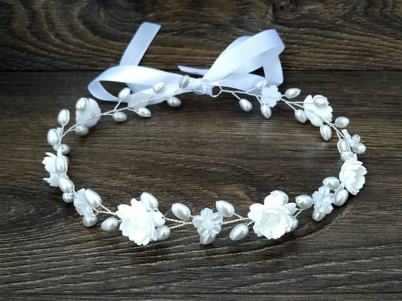 May include: A white floral crown with pearl accents and a white satin ribbon. The crown is made of wire and is decorated with small white flowers and pearls.