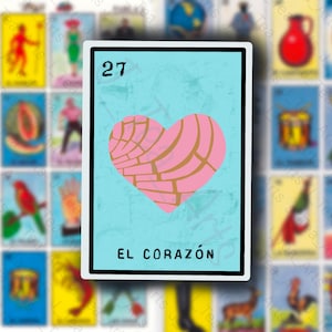 May include: A blue and white card with the number 27 printed on it. The card features a pink heart-shaped pastry with a gold design. The text "EL CORAZÓN" is printed below the pastry.