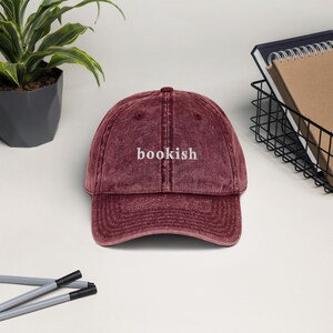 May include: A maroon baseball cap with the word "bookish" embroidered in white. The cap has a curved brim and a vintage, worn look. The hat is a stylish accessory for book lovers.