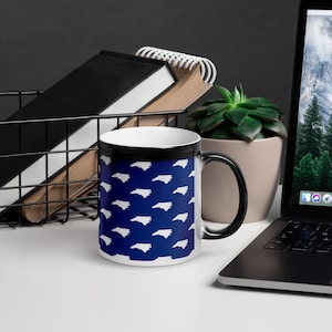 May include: A black and white magic mug with a blue background. The mug features a repeating pattern of the outline of the state of North Carolina.