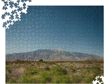 Può includere: Un puzzle completo raffigurante un paesaggio montano sotto un cielo blu. Il puzzle è quasi interamente assemblato, con alcuni pezzi sparsi sui bordi. La scena comprende vegetazione verde e marrone in primo piano.