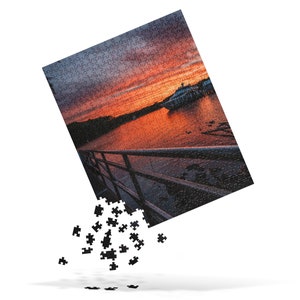 May include: A jigsaw puzzle with a sunset over a body of water. The puzzle is partially assembled, with some pieces missing. The image shows a boat in the water and a railing in the foreground.