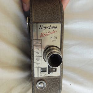 May include: A vintage Keystone Pathfinder K-29 8mm film camera with a silver lens and a grey body. The camera has a dial for adjusting the exposure settings for colour film.