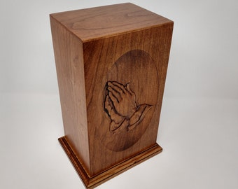 May include: A wooden cremation urn with a carved design of praying hands on the front. The urn is a rich brown color and has a simple, elegant design.
