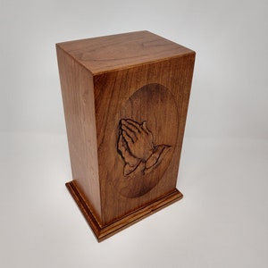 May include: A wooden cremation urn with a carved design of praying hands on the front. The urn is a rich brown color and has a simple, elegant design.