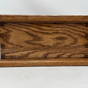 May include: A wooden box with a natural wood finish. The box has a raised lid and a base with a slightly rounded edge. The wood grain is visible on the surface of the box.