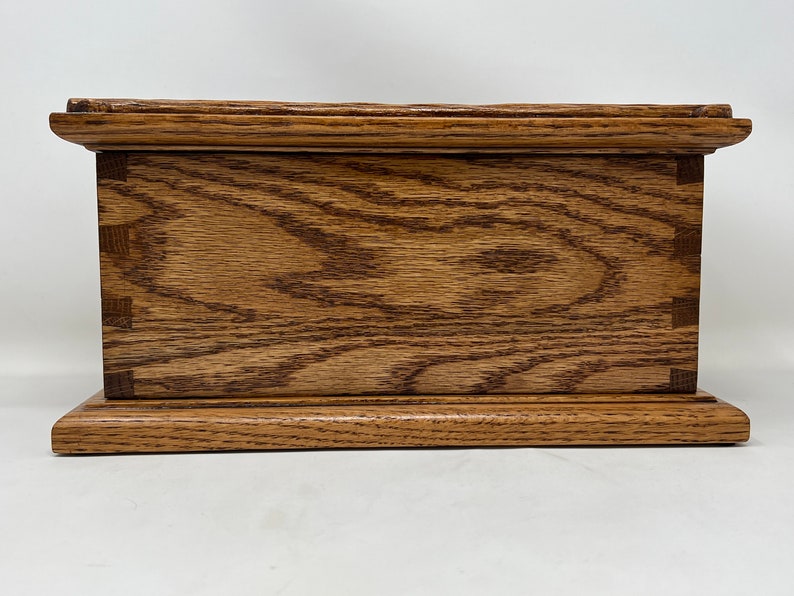 May include: A wooden box with a natural wood finish. The box has a raised lid and a base with a slightly rounded edge. The wood grain is visible on the surface of the box.