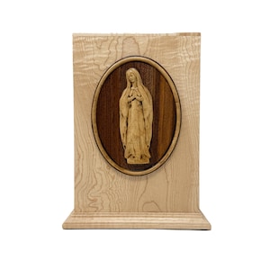 May include: A wooden religious plaque featuring a carved image of a saint within an oval frame. The frame is set against a light-colored wooden background. The carving is detailed and the wood has a natural grain.
