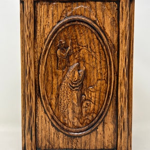 May include: A wooden box with a carved design of a fisherman holding a fish. The box is dark brown and has a rustic look.