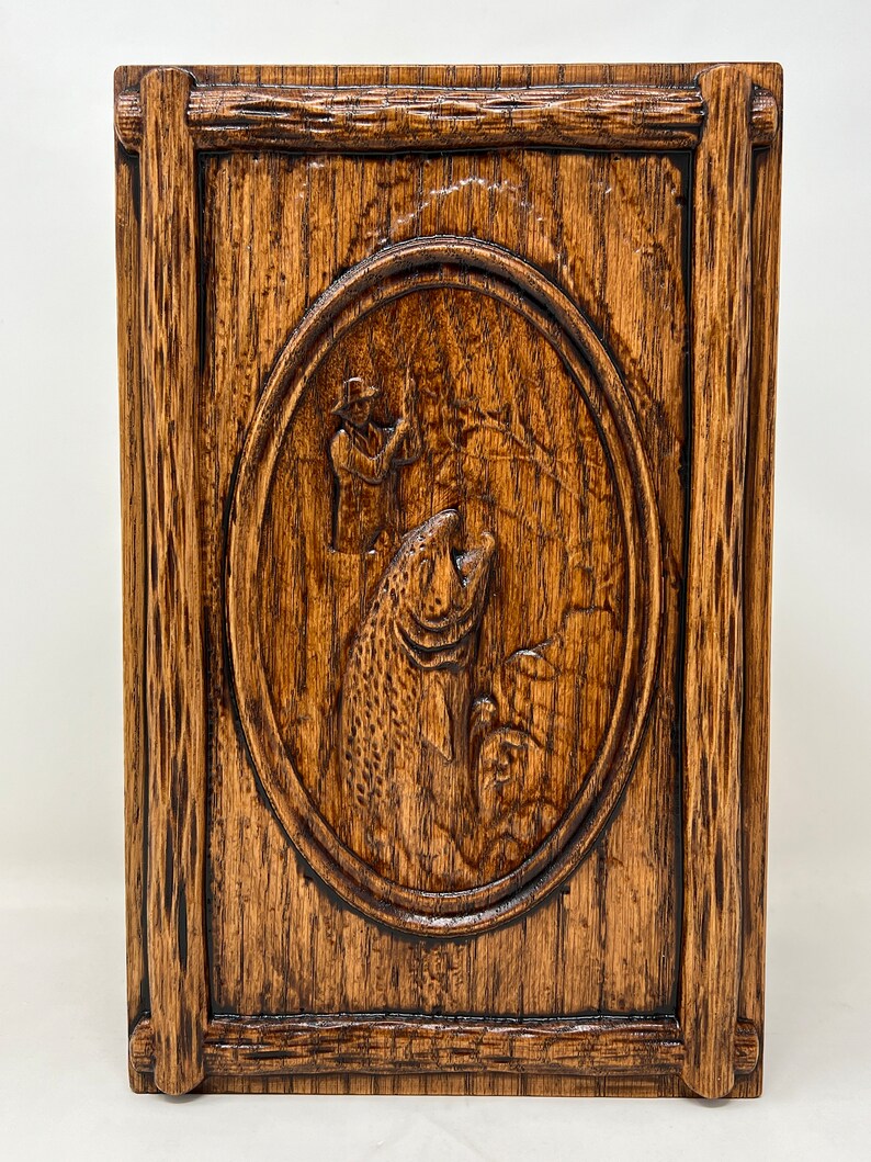 May include: A wooden box with a carved design of a fisherman holding a fish. The box is dark brown and has a rustic look.