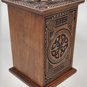 May include: A wooden box with intricate Celtic knotwork carvings. The box is made of dark wood and has a square shape. The top of the box features a Celtic knot design in the center.